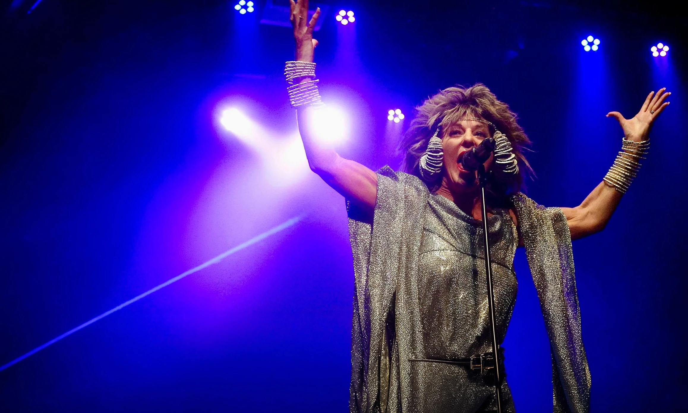 Julie Nevada performing as Tina Turner with her arms raised beneath blue stage lights